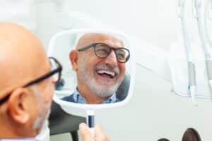 Happy senior patient admiring his new smile after dental implant treatment, holding a mirror at the dental clinic.