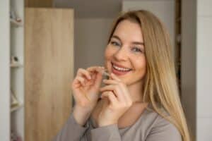 Cheerful blonde woman with freckles smiling warmly while holding a clear orthodontic aligner, representing modern dental care and teeth straightening