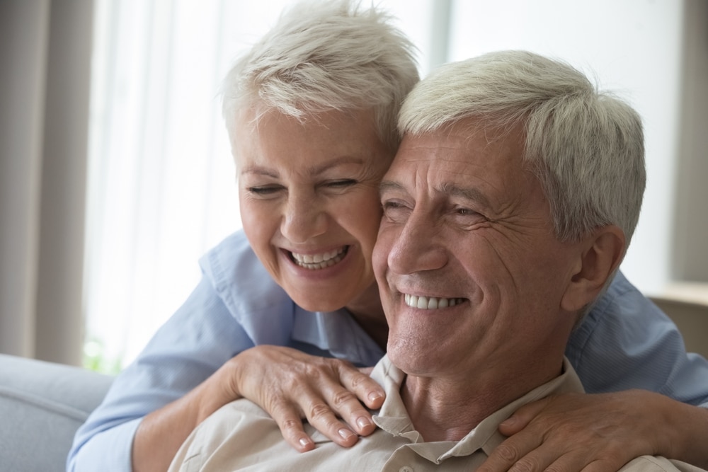 Affectionate middle-aged wife embracing husband from behind, smiling while he shows his teeth after dental bonding treatment.