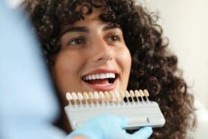 Dentist checking young woman’s teeth color in clinic during consultation for dental veneers and cosmetic dentistry