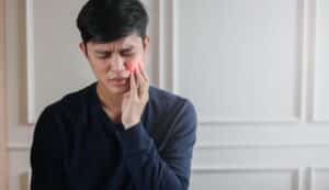 Close-up of young man frowning and massaging his cheek due to TMJ pain, concept of jaw muscle tension and treatment for unhealthy lifestyle habits.