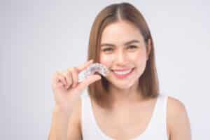 Woman smiling and showing off her straight, white teeth while holding up her clear aligners.