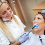 Beautiful young smiling woman at visit in the dentist office and dentist finishing repair tooth with dental curing UV lamp.
